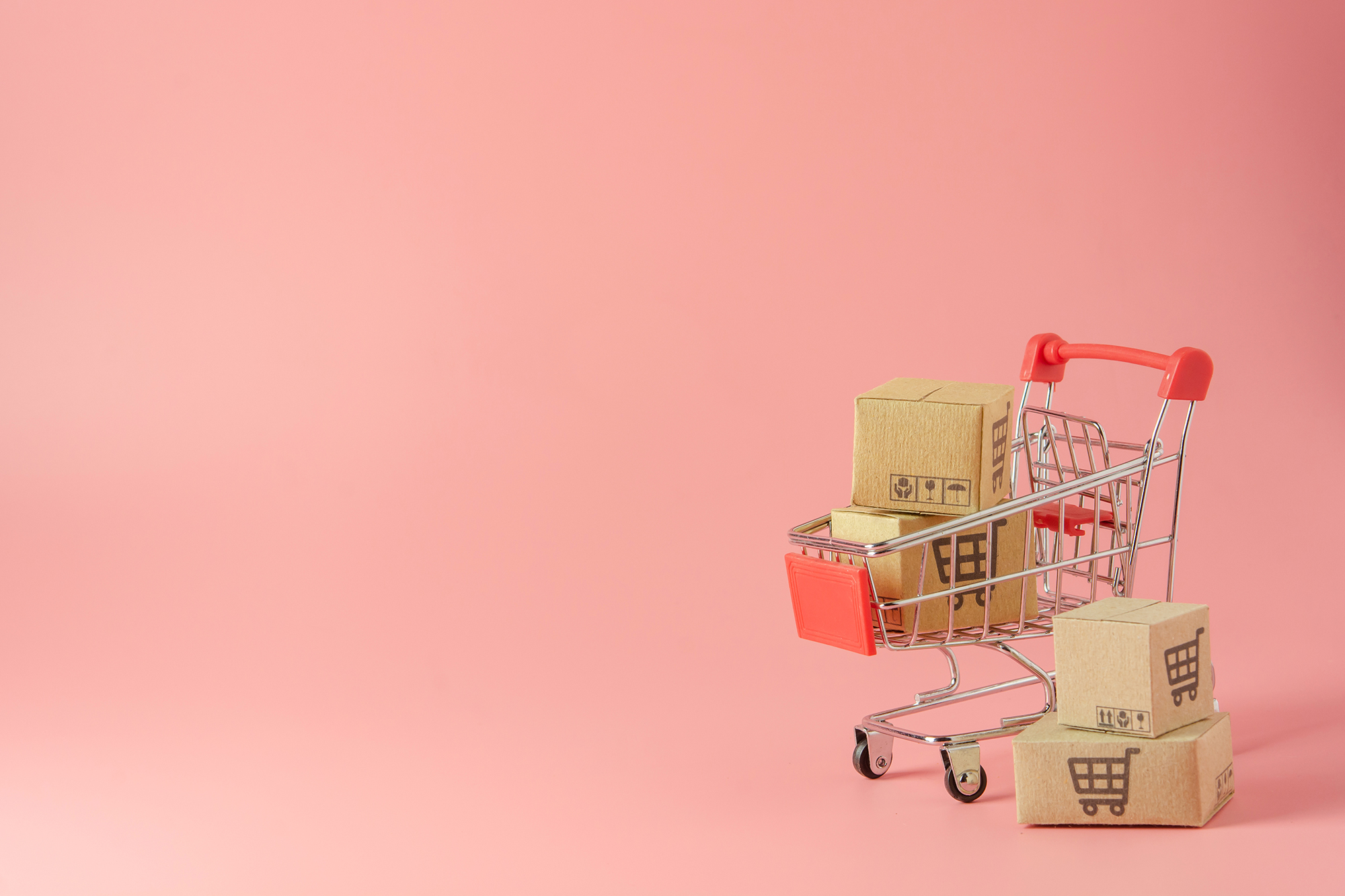 Image représentant des cartons dans un caddie de supermarché miniature. Sur un fond rose.